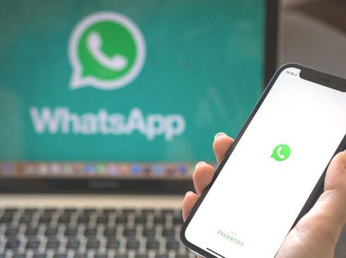 Pessoa com um celular na mão, em que aparece o ícone do WhatsApp. Ao fundo, um computador de mesa, com o símbolo da rede social também aparecendo na tela.