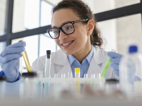 Mulher de óculos, jaleco branco e luvas azuis manipula tubos de ensaio em ambiente de pesquisa/laboratório.