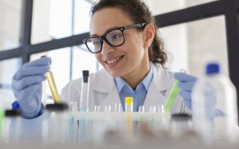 Mulher de óculos, jaleco branco e luvas azuis manipula tubos de ensaio em ambiente de pesquisa/laboratório.