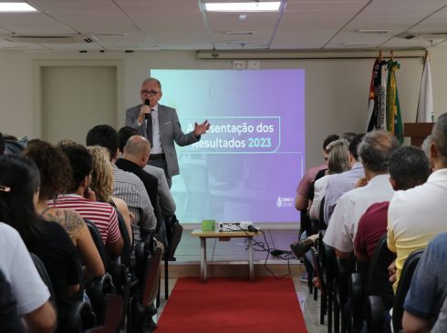 Um homem, de terno e gravata, segura um microfone na mão, no palco de um auditório. Ele está sendo observado pela plateia, homens e mulheres sentados em cadeiras.