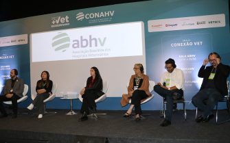 Seis pessoas sentadas lado a lado, em cima de um palco, cada uma em uma cadeira branca atrás delas, o logo da ABHV(Associação Brasileira dos Hospitais Veterinários).