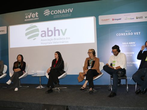 Seis pessoas sentadas lado a lado, em cima de um palco, cada uma em uma cadeira branca atrás delas, o logo da ABHV(Associação Brasileira dos Hospitais Veterinários).