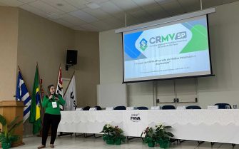 Mulher vestindo blusa verde e calça preta falando no microfone durante Simpósio Paulista de Leishmaniose Visceral.