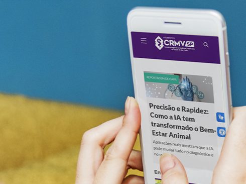 Mão feminina branca segurando celular branco com a página principal do CRMV-SP.