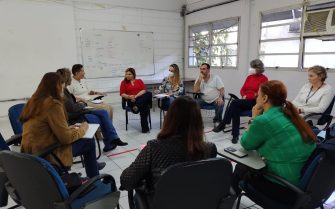 Na imagem há nove pessoas sentadas em círculos, em uma sala de reunião, com uma lousa ao fundo.