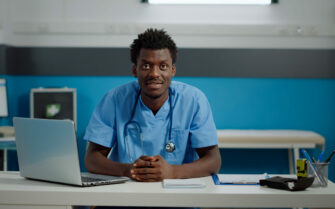 Médico-veterinário negro está sentado atrás de uma mesa com computador, olhando e sorrindo para a câmera.