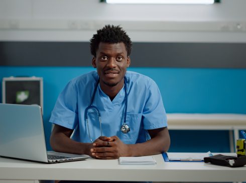 Médico-veterinário negro está sentado atrás de uma mesa com computador, olhando e sorrindo para a câmera.
