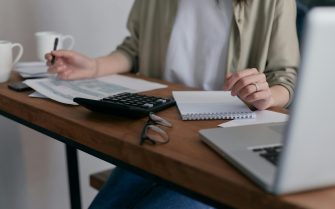 Pessoa sentada atrás de uma mesa de madeira em que estão colocados papéis, uma calculadora e um notebook. Ela segura uma caneta em uma das mãos e seu rosto não aparece na fotografia.