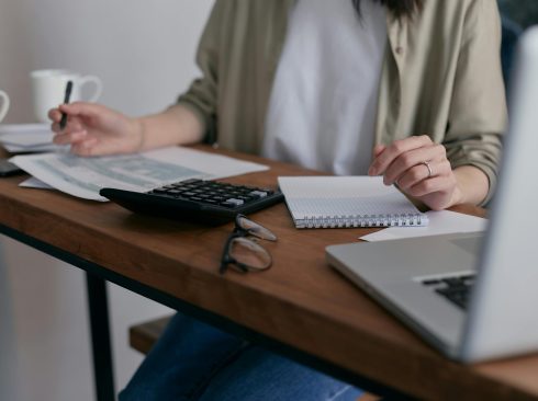 Pessoa sentada atrás de uma mesa de madeira em que estão colocados papéis, uma calculadora e um notebook. Ela segura uma caneta em uma das mãos e seu rosto não aparece na fotografia.