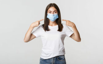 Covid-19, health and social distancing concept. Attractive brunette girl in medical mask pointing finger at face, white background