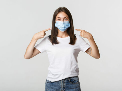 Covid-19, health and social distancing concept. Attractive brunette girl in medical mask pointing finger at face, white background