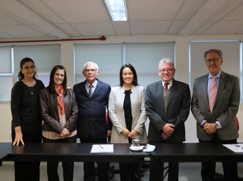 Na imagem há seis pessoas de pé sorrindo, em frente a uma mesa,sendo três mulheres e três homens.