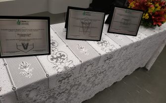 Mesa com toalha branca contendo 3 placas de homenagem e flores.