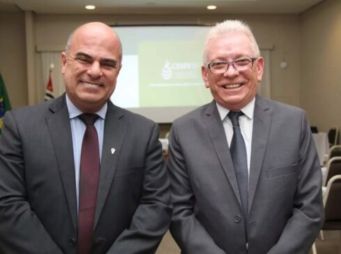 Presidente e vice-presidente do CRMV-SP posam juntos vestindo terno e gravata. Ambos estão sorrindo e dispostos em frente a um telão com a logomarca do CRMV-SP sob um fundo verde.