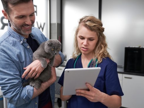 A foto mostra uma médica-veterinária segurando um tablet. Ao lado dela há um homem com um gato no colo.