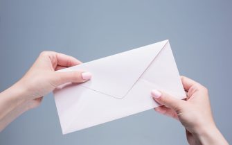 Imagem mostra duas mãos femininas de pele clara, segurando um envelope branco fechado.