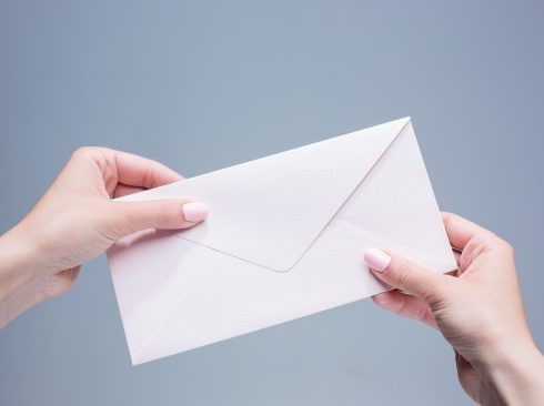 Imagem mostra duas mãos femininas de pele clara, segurando um envelope branco fechado.