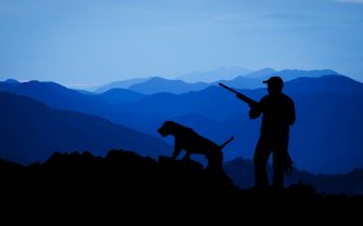 A imagem traz a sombra de um caçador com uma arma nas mãos e um cão em uma paisagem natural de campo e montanhas. Há diferentes tons e sobretons de azul compondo a cena.