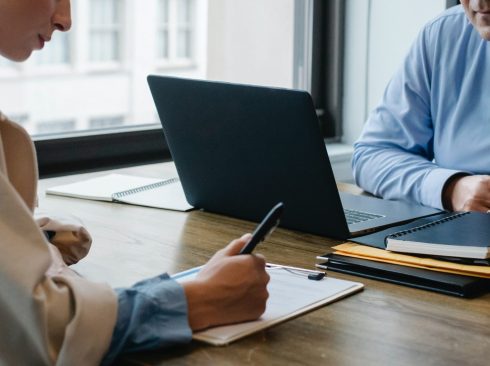 Na imagem aparecem dois profissionais, um homem e uma mulher, sentados em volta de uma mesa de trabalho com materiais para anotação e um notebook.