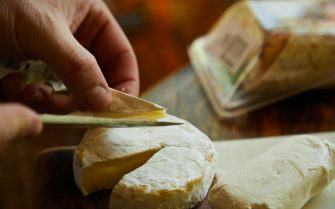Mão masculina utiliza uma faca para cortar uma fatia de um queijo amarelado curado