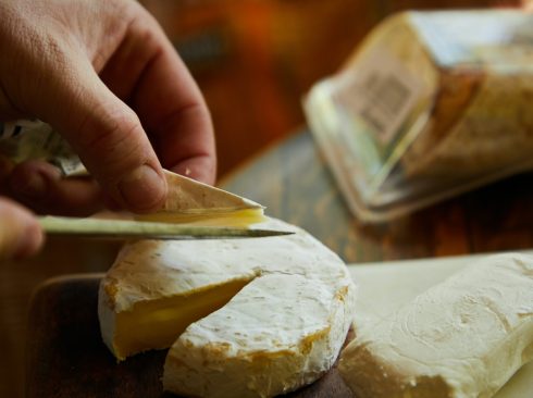 Mão masculina utiliza uma faca para cortar uma fatia de um queijo amarelado curado