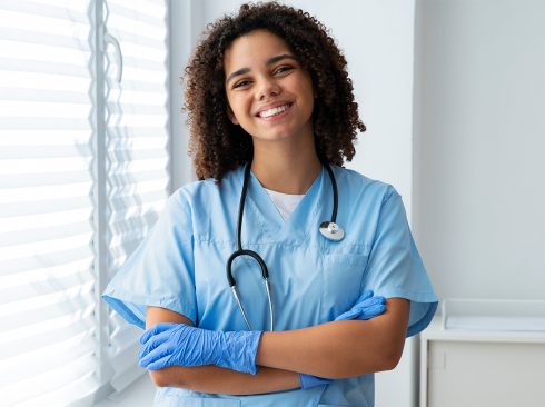 Mulher jovem, negra de cabelos cacheados na altura dos ombros, sorrindo para foto. Ela veste uniforme e luvas cirúrgicas azuis e tem um estetoscópio no pescoço.