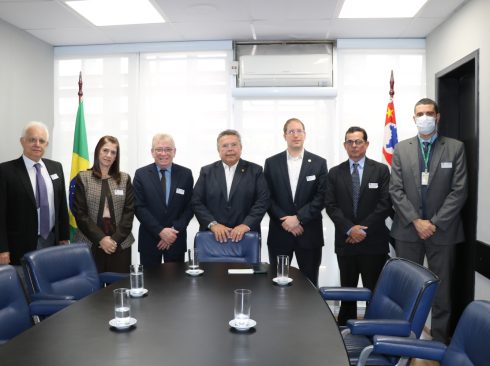 Na imagem há sete pessoas em pé, em frente a uma mesa grande de reunião, sendo uma mulher e seis homens.