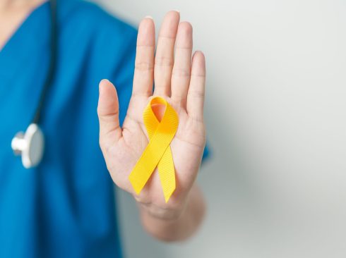 A imagem mostra o corpo de uma médica-veterinária branca, em pé, vestindo uniforme azul e com o estetoscópio no pescoço com o braço direito esticado e a mão direita em riste com um laço amarelo da campanha setembro amarelo na mão direita.
