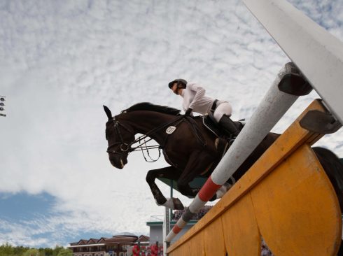 A imagem mostra de baixo para cima, um cavalo de salto com pelagem marrom escura pulando um obstáculo durante competição, enquanto é montado por uma jóquei mulher jovem vestindo uniforme branco.