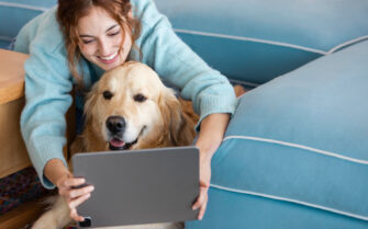 A foto mostra uma mulher com seu cachorro à sua frente. Ela está sorrindo e segura um tablet, à frente dele e do cachorro.