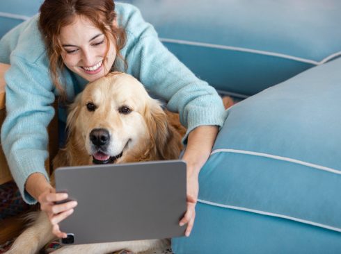 A foto mostra uma mulher com seu cachorro à sua frente. Ela está sorrindo e segura um tablet, à frente dele e do cachorro.