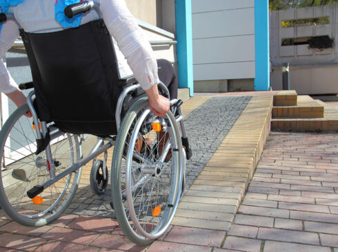 Foto mostra pessoa em cadeira de rodas, subindo uma rampa que dá acesso a um estabelecimento.