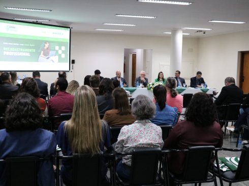 Na imagem aparece um auditório lotado de profissionais. Eles estão de costas para a câmera, sentados em cadeiras e dispostos de frente para a diretoria do Conselho, que está a frente sentada atrás de uma mesa com toalha branca e arranjo de flores. Na sala há ainda um telão em que se destaca o site do CRMV-SP com banner sobre a campanha de recadastramento.