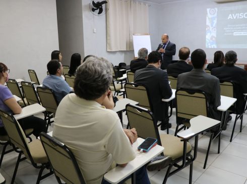 Foto mostra um auditório com carteiras escolares e pessoas nelas sentadas. A frente está o vice-presidente do CRMV-SP falando ao microfone. Ao fundo um telão com aviso sobre as restrições inerentes ao período eleitoral.