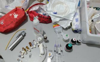 Mesa com ampolas abertas, seringas agulhadas com conteúdo não identificado, medicações de uso controlado, e equipamentos de uso cirúrgico.