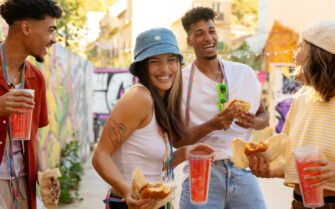 A foto mostra duas mulheres e dois homens brincando o carnaval. Eles estão alegres, usam acessórios coloridos e seguram bebidas de cor laranja e sanduíches tipo hambúrguer, nas mãos.