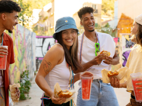 A foto mostra duas mulheres e dois homens brincando o carnaval. Eles estão alegres, usam acessórios coloridos e seguram bebidas de cor laranja e sanduíches tipo hambúrguer, nas mãos.