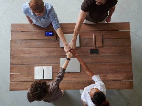 A imagem mostra quatro pessoas juntando as mãos, representando a união, em torno de uma mesa retangular de madeira.