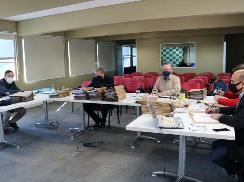 Membros da Tomada de Contas estão de máscara, sentados ao redor de mesas com computadores e pastas com processos