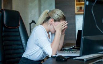 Mulher desesperada com as mãos no rosto, em sua mesa de trabalho, em frente a um computador.