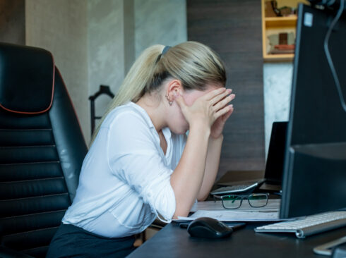 Mulher desesperada com as mãos no rosto, em sua mesa de trabalho, em frente a um computador.