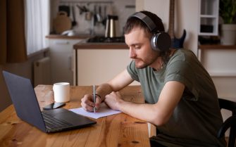 Rapaz em frente a um notebook, apoiando em uma mesa e fazendo anotações em um papel. Ele está sentado, usa fones de ouvido e camisa verde.
