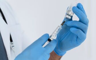 Imagem mostra profissional da saúde segurando uma seringa que está extraindo líquido de um pequeno frasco.