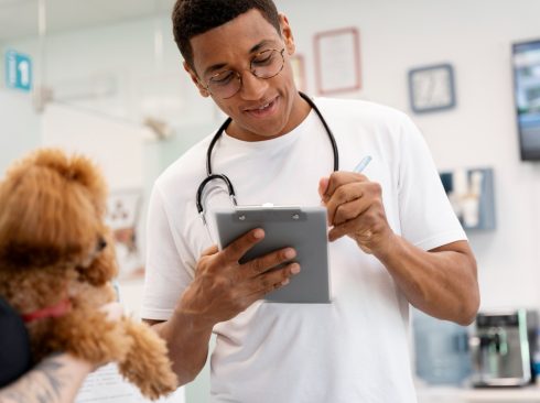 médico-veterinário em pé usando óculos e vestindo blusa branca, escreve no prontuário enquanto mulher segura cachorro peludo de porte médio.