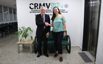 Presidente do CRMV-SP, Odemilson Donizete Mossero, parabeniza a presidente eleita para a gestão 2024-2027 do Regional. Eles olham para a câmera e fazem um cumprimento de mão, sorrindo, em frente a logomarca do Conselho.