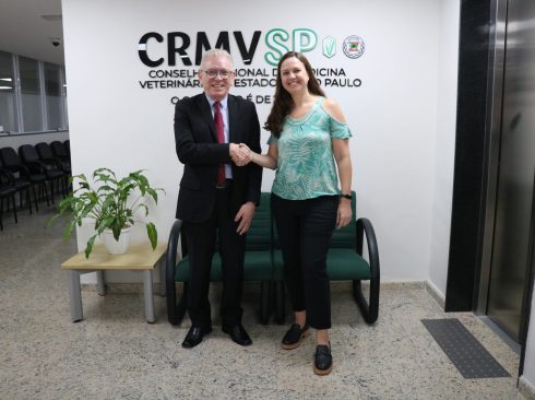 Presidente do CRMV-SP, Odemilson Donizete Mossero, parabeniza a presidente eleita para a gestão 2024-2027 do Regional. Eles olham para a câmera e fazem um cumprimento de mão, sorrindo, em frente a logomarca do Conselho.