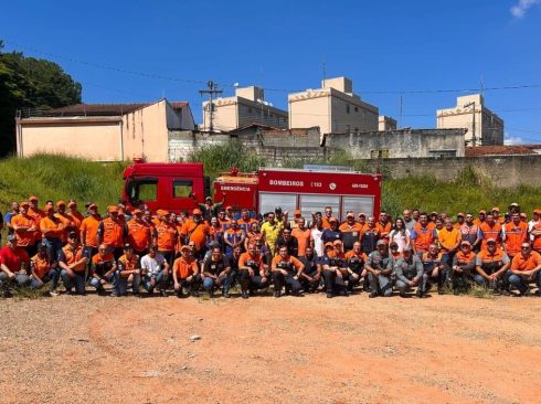 Equipe formada por profissionais de diversos setores, entre mulheres e homens posam em três fileiras em frente a um carro do Corpo de Bombeiros