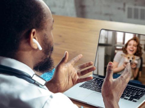 Médico-veterinário está sentando em frente ao notebook fazendo uma teleconsulta. Na tela, uma tutora sorrindo com o seu cão.