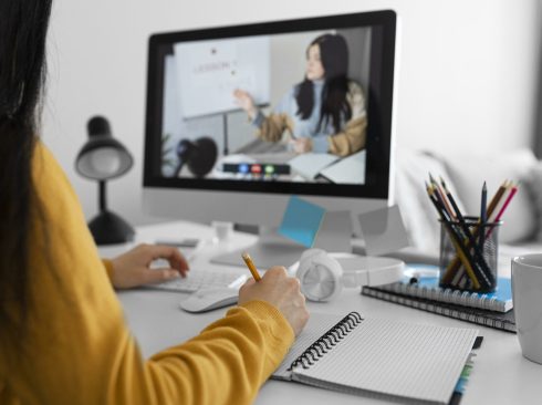 Mulher em frente ao computador, com caderno na mesa e lápis em uma das mãos e teclado na outra mão, assistindo aula