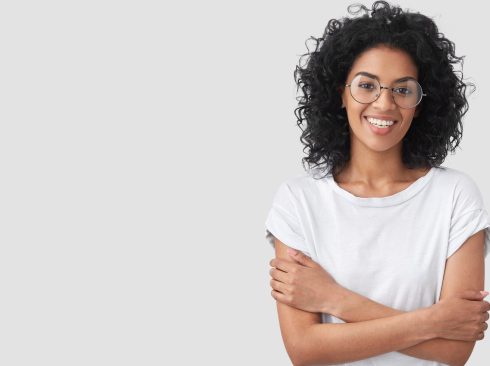 Positive dark skinned female has toothy smile, shy expression, keeps arms folded, looks happily at camera, glad to have peasant talk with friend, isolated on white background with blank space on left